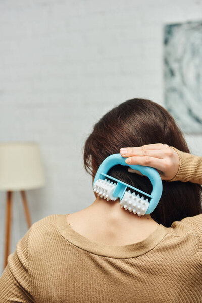 Вид сзади на молодую женщину в непринужденном коричневом джампере, держащую массажер и делающую самомассаж спины в размытой гостиной, поддержку лимфатической системы и домашний массаж, снятие напряжения