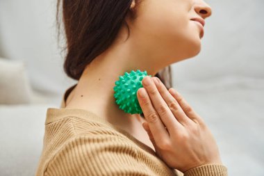 Cropped view of brunette woman in casual jumper massaging lymphatic system and nodes on neck with manual massage ball at home, lymphatic system support and home-based massage