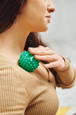 Cropped view of brunette woman in brown jumper massaging lymphatic system on chest with manual massage ball at home, self-care ritual and holistic wellness practices concept, balancing energy