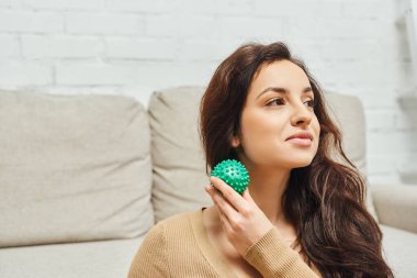 Young brunette woman in jumper holding manual massage ball and looking away while sitting near couch at home, home-based massage and holistic wellness practices concept, balancing energy clipart