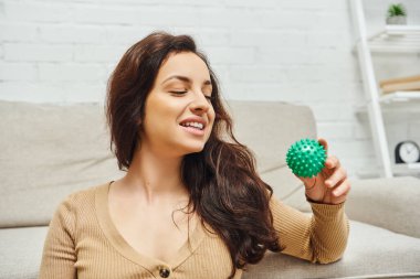 Cheerful young brunette woman in casual jumper looking at manual massage ball while sitting near couch at home, home-based massage and holistic wellness practices concept, balancing energy clipart