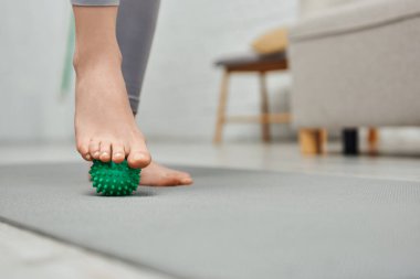 Cropped view of woman massaging foot with manual massage ball and standing on fitness mat at home, body relaxation and holistic wellness practices, balancing energy