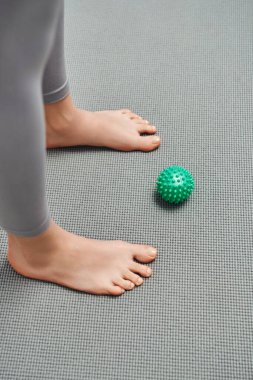 Çıplak ayaklı bir kadının evdeki spor minderinin yanında duran manuel masaj topunun en üst görüntüsü, vücut gevşemesi ve bütünsel sağlık uygulamaları, enerji dengeleme.