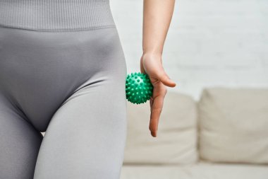 Cropped view of young woman in sportswear massaging hip with manual massage ball in blurred living room, body relaxation and holistic wellness practices, tension relief clipart