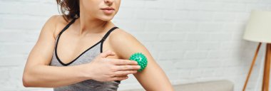 Cropped view of young woman in sportswear massaging arm with manual massage ball while standing at home, balancing energy and holistic healing concept, banner, myofascial release clipart