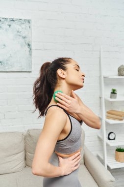 Spor kıyafetli genç esmer kadın masaj topuyla boyna masaj yapıyor ve oturma odasında duruyor. Enerji ve bütünsel iyileşme kavramını dengeliyor.