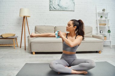 Spor giyim bölümündeki genç esmer kadın masajcı koluyla evde spor minderinin üzerinde otururken enerji ve bütünsel iyileşme kavramını dengeliyor, miyofasyal rahatlama.