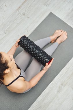 Aktif kıyafetli, tekerlekli sandalyede oturan ve oturma odasındaki spor minderinde oturan çıplak ayaklı genç bir kadının genel bakış açısı, ev tabanlı masaj ve bütünsel uygulamalar konsepti, miyofassiyonel rahatlama.