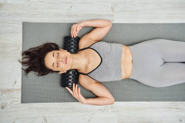 Spor kıyafetli genç esmer kadının boynundaki paten masajını evde spor minderinin üzerinde yatarken, ev yapımı masaj ve bütünsel uygulama konsepti, miyofasiyal rahatlama