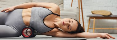 Genç ve rahat esmer kadın spor kıyafetleri içinde masaj masörüyle vücuduna masaj yapıyor ve oturma odasındaki spor paspasına uzanıyor, kendine bakım ve refah kavramına odaklanıyor, afiş. 