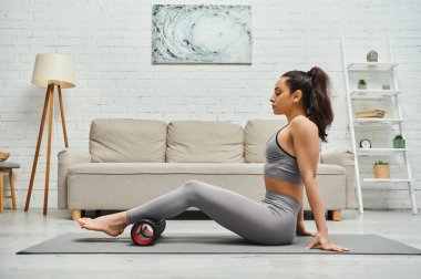 Rahatlamış esmer kadın spor kıyafetleri içinde bacakları tekerlekli masajla ovuyor ve evdeki oturma odasında mindere oturuyor sağlıklı lenfatik sistem kavramını koruyor, miyofasiyal rahatlama sağlıyor.