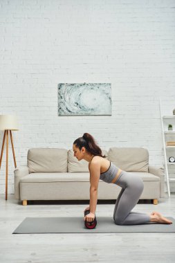 Spor kıyafetli genç esmer kadın, elinde modern masaj silindiri tutarken ve evdeki spor paspası üzerinde çalışırken, sakinlik hissi ve rahatlama konseptini desteklerken yan görünüm.