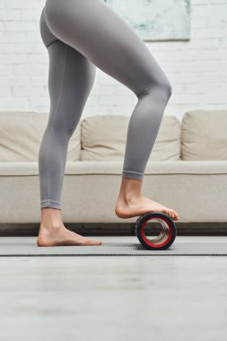 Aktif giyinen, kendine masaj yapan, modern tekerlekli sandalyede spor paspası takan, ev konseptinde lenf akışını ve sağlığını destekleyen çıplak ayaklı genç bir kadın görüntüsü.