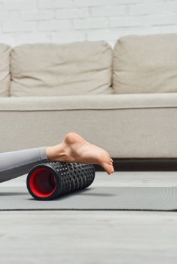 Çıplak ayaklı bir kadının, evdeki oturma odasının yakınındaki fitness minderinde uzanırken modern masajcıyla bacağa masaj yapması, ev konseptinde lenf akışını ve sağlığını teşvik etmesi, gerilim atağı.