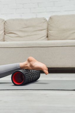 Cropped view of barefoot woman massaging leg with modern roller massager while lying on fitness mat near living room at home, promoting lymph flow and wellness at home concept, tension relief clipart