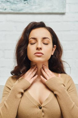 Portrait of young brunette woman in brown jumper checking thyroid gland on neck while sitting with closed eyes on couch in living room at home, focus on self-care and well-being concept 