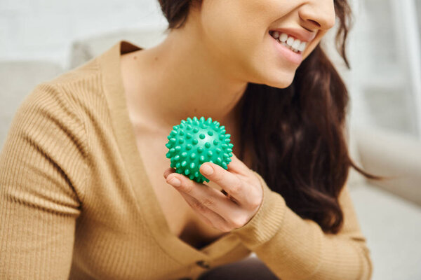 Cropped view of cheerful young brunette woman in brown jumper holding manual massage ball in blurred living room, body relaxation and holistic wellness practices, balancing energy