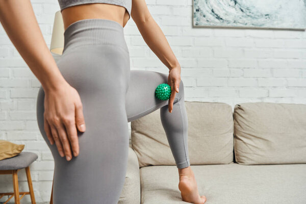 Cropped view of woman in sportswear massaging muscle on leg with manual massage ball near couch at home, holistic wellness practices and body relaxation concept, tension relief