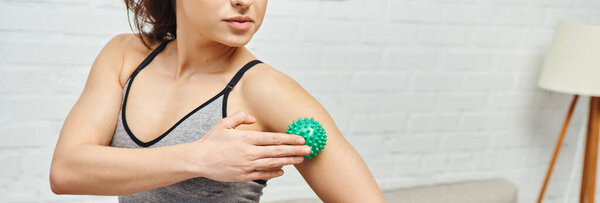 Cropped view of young woman in sportswear massaging arm with manual massage ball while standing at home, balancing energy and holistic healing concept, banner, myofascial release