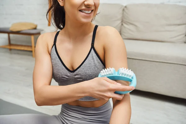 stock image Cropped view of smiling young woman in sportswear massaging arm with handled massager in blurred living rom at home, balancing energy and holistic healing concept, myofascial release