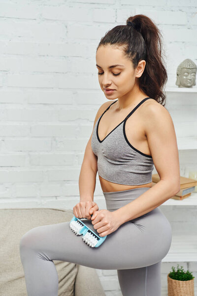 Молодая женщина в активной одежде делает лимфатическую циркуляцию на ноге с ручным массажером рядом с диваном в гостиной дома, домашний массаж и целостные практики концепции, миофасциальное освобождение