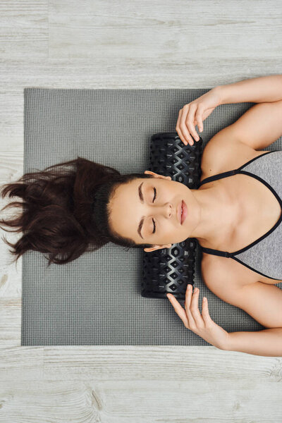 Top view of young relaxed brunette woman in sportswear using roller massager on neck while lying on fitness mat at home, home-based massage and holistic practices concept, myofascial release