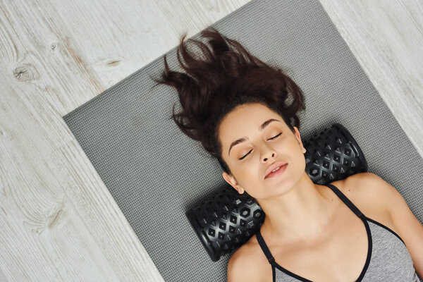 Top view of relaxed young brunette woman with closed eyes using roller massager on neck while lying on fitness mat at home, home-based massage and holistic practices concept, myofascial release