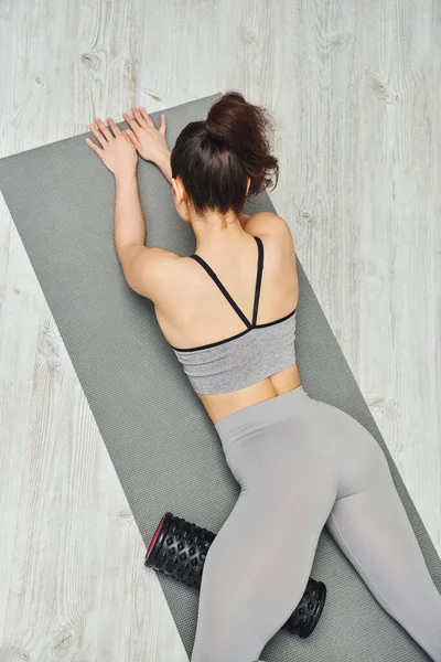 stock image Back view of young brunette woman in sportswear massaging leg with roller massager and lying on fitness mat on floor at home, focus on self-care and well-being concept, tension relief