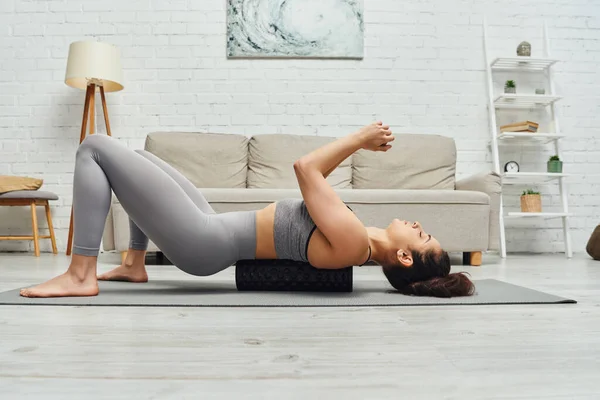 stock image Side view of young brunette woman in sportswear lying on roller massager and fitness mat during self-massage in living room at home, sense of tranquility, relaxation concept, tension relief