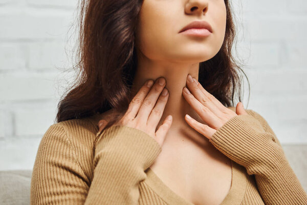 Cropped view of young brunette woman in brown jumper checking thyroid gland on neck while sitting on blurred couch in living room at home, focus on self-care and well-being concept 