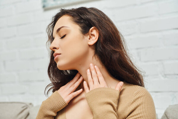 Young brunette woman in casual clothes touching neck while checking thyroid gland and sitting on couch in living room at home, focus on self-care and well-being concept, tension relief