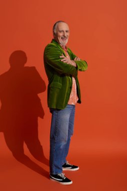 Yakışıklı ve sakallı, kolları bağlı, kırmızı turuncu arka planda kameraya gülümseyen yeşil kadife ceketli, mavi kot pantolonlu, şık bir yaşlanma konsepti olan yaşlı bir adam.