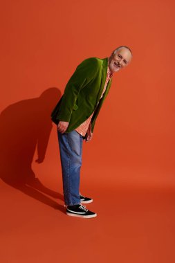 full length of joyful and bearded senior man in trendy casual clothes standing and looking at camera on red orange background, green velour blazer, blue denim jeans, happy aging concept clipart