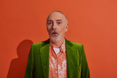 surprised and impressed senior man with bulging eyes looking at camera on red orange background, grey hair and beard, green velour blazer and trendy shirt, personal style and positive aging concept clipart