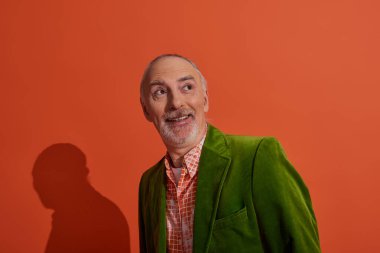 joyful and surprised senior man with grey hair and beard smiling and looking away on red orange background, fashionable casual clothes, green velour blazer, trendy shirt, positive aging concept clipart