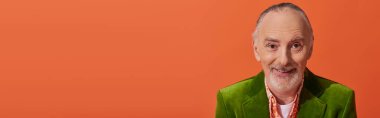 portrait of trendy and cheerful senior model with grey hair and beard, wearing green velour blazer and smiling at camera on vibrant orange background, fashion and age concept, banner with copy space clipart