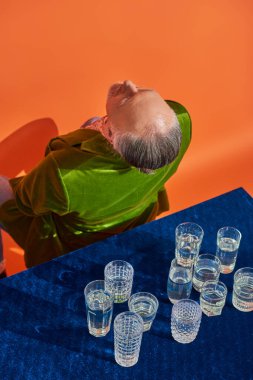 Yeşil kadife ceketli üst düzey bir adam, mavi kadife kumaşlı ve turuncu arka planda bir bardak suyla masada oturuyor. Yaşlanan nüfus, sembolizm, hayat doluluk kavramı.