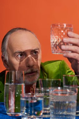 yeşil kadife ceketli yaşlı ve düşünceli bir adam. Masanın yanında saf su dolu bardağa bakıyor. Turuncu arka planda mavi kumaş, yaşlı nüfus, sembolizm, yaşam doluluk kavramı.