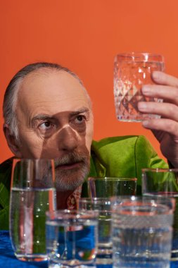 senior and thoughtful grey haired man in green velvet blazer looking at glass full of pure water near table with blue cloth on orange background, aging population, symbolism, life fullness concept clipart