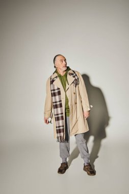 full length of discouraged and thoughtful senior man in stylish casual clothes looking away on grey background with shadow, beige trench coat, green hoodie, plaid blazer, age positive concept clipart