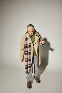 Gri arka planda, gölgeli, bej renkli trençkot, ekose atkı, yeşil kapüşonlu, moda ve yaş kavramına sahip modaya uygun giyinmiş bir şekilde dikilen yaşlı sakallı bir adam. 