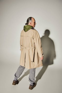 back view of senior model in stylish casual clothes standing on grey background with shadow, aged and grey haired man in beige trench coat and green hoodie, trendy lifestyle concept clipart
