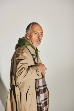 Gri saçlı, sakallı ve düşünceli son sınıf öğrencisi gri arka planda şık kıyafetlerle poz veriyor, bej rengi trençkot, yeşil kapüşonlu, ekose atkı, moda ve yaş, yaşlanan nüfus kavramı.