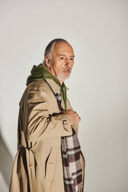 grey haired, bearded and pensive senior man posing in stylish casual clothes on grey background, beige trench coat, green hoodie, plaid scarf, fashion and age, aging population concept clipart