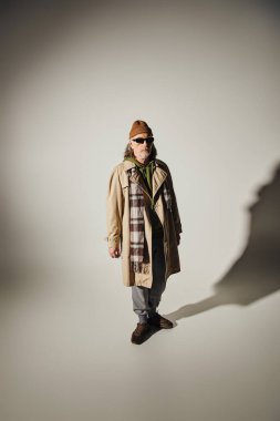 fashion and age concept, full length of hipster style senior man in beanie hat, dark sunglasses, beige trench coat and plaid scarf standing on grey background with shadow and copy space clipart