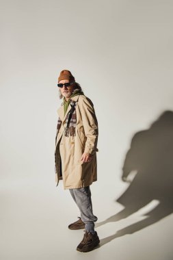 full length of fashionable senior model in dark sunglasses, beanie hat, beige trench coat and plaid scarf standing on grey background with shadow, positive and trendy aging concept clipart