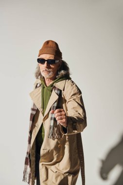 Koyu renk güneş gözlüklü ve hippi tarzı giyinen kendinden emin bir adam. Yumruğunu sıkmış ve gri arka planda kameraya bakıyor. Şapka, bej rengi yağmurluk, modaya uygun yaşlanma konsepti.