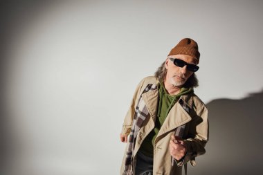 Yaşlı ve sakallı adam, koyu güneş gözlüklü, bere şapkalı, ekose atkı ve bej renkli trençkotlu gri arka plandaki kameraya bakıyor. Gölge ve fotokopi alanı, hipster tarzı, moda ve yaş kavramı.