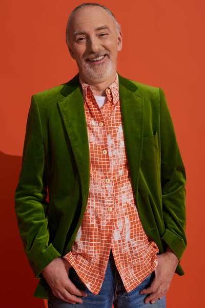 carefree, grey haired and fashionable senior man in green velour blazer and trendy shirt standing with thumbs in pockets and smiling at camera on red orange background, happy aging concept