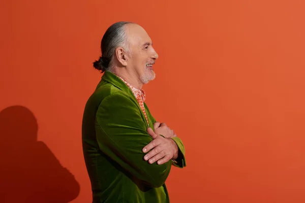 stock image side view of senior male model, optimistic man posing with folded arms and smiling on red orange background, green velour blazer, grey hair and beard, positive and fashionable aging concept